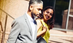 Hombre y mujer de negocios, sonrientes caminando afuera de unas oficinas bajo el sol.