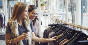 Dos mujeres riendo, con café en la mano, y tocando y mirando ropa colgada en perchas, en una tienda al por menor de un centro comercial.
