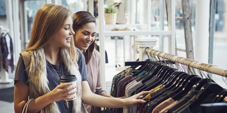 Dos mujeres riendo, con café en la mano, y tocando y mirando ropa colgada en perchas, en una tienda al por menor de un centro comercial.