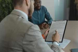 Two men in office attire review a document on a clipboard at a round table, discussing terms.