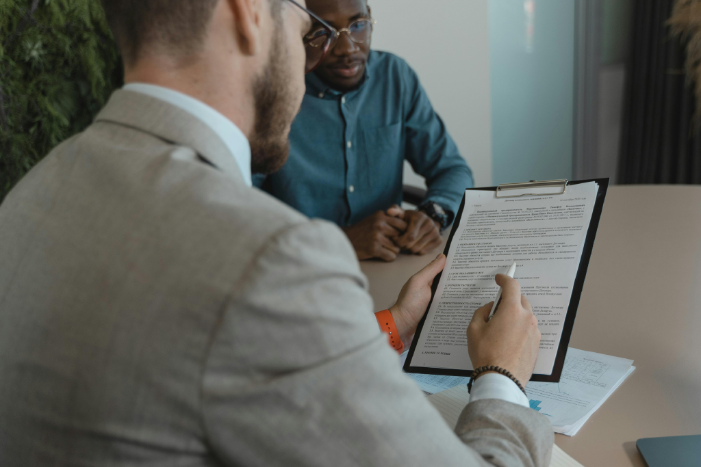 Two men in office attire review a document on a clipboard at a round table, discussing terms.