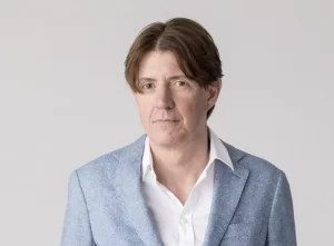 Man in a light blue blazer and white shirt poses for a head-and-shoulders portrait against a plain light background.