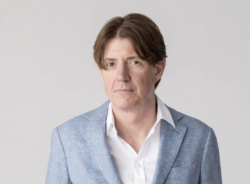Man in a light blue blazer and white shirt poses for a head-and-shoulders portrait against a plain light background.