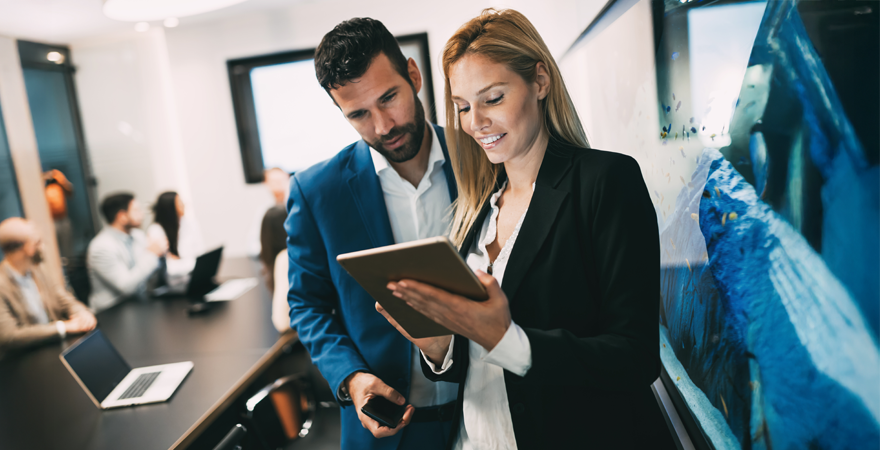 Two professionals in business attire review a tablet together during a meeting in a modern office with colleagues at a conference table in the background, near a large wall display.