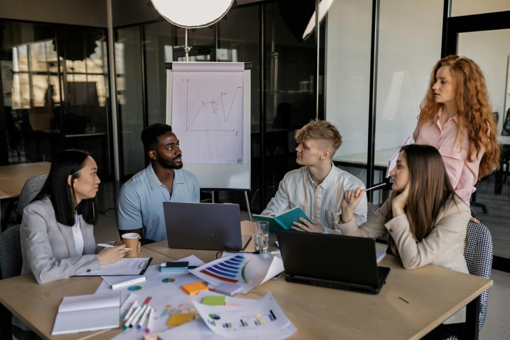 Team of five in a modern office around a table with laptops and charts, discussing a graph on a flip chart.