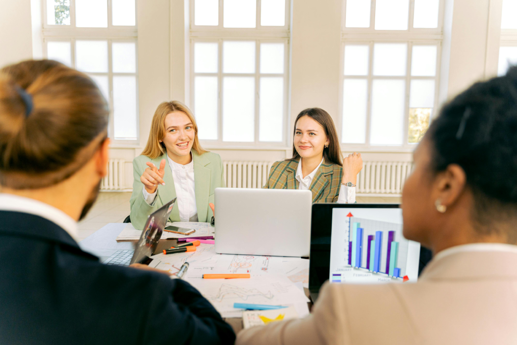 Four professionals in a bright conference room sit around a table with laptops, discussing charts and notes.