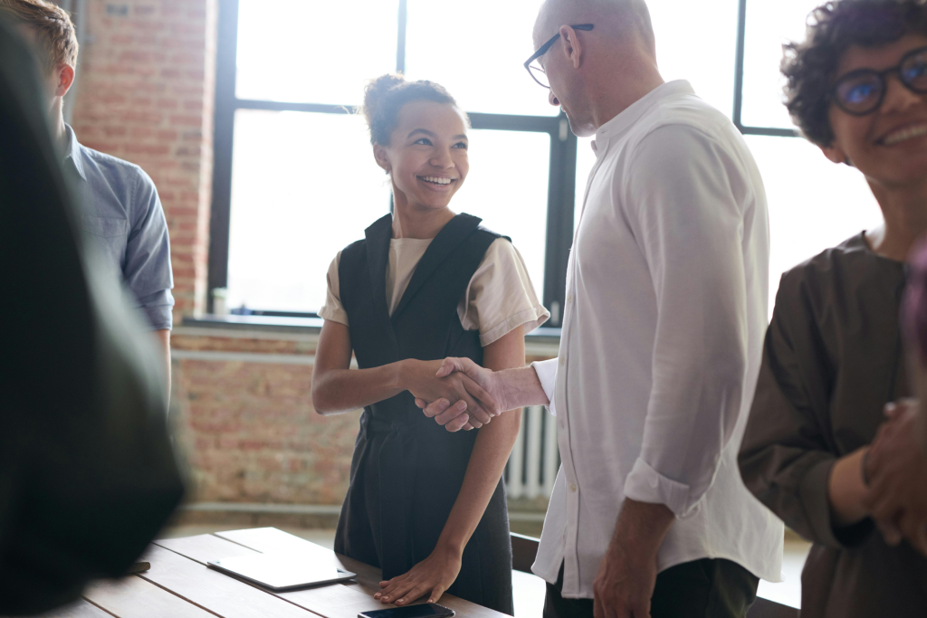 Two colleagues shake hands at a sunlit office table, the woman smiling as they greet; others chat in the background.