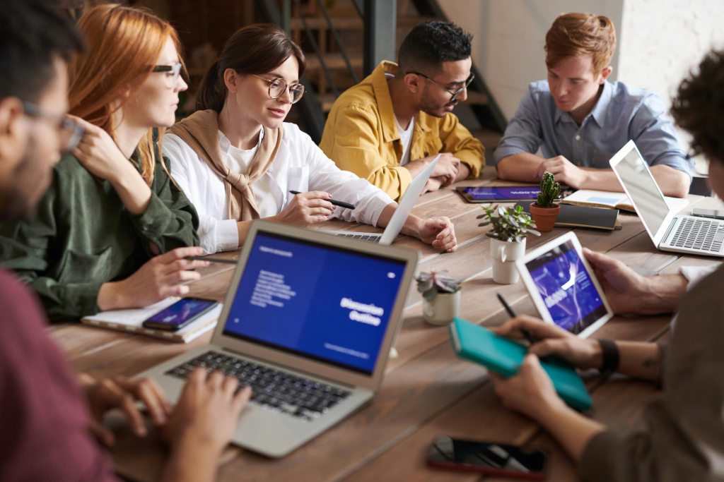Group of diverse coworkers collaborating around a wooden table with laptops, tablets, and notebooks.