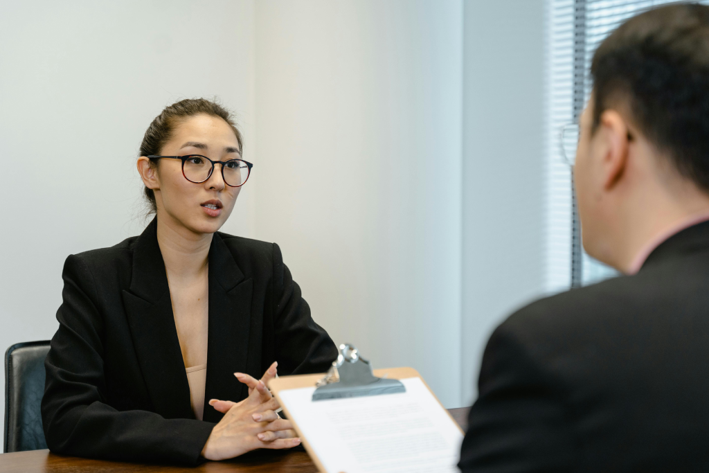 Asian woman in a black blazer speaks during a meeting across a table in an office.