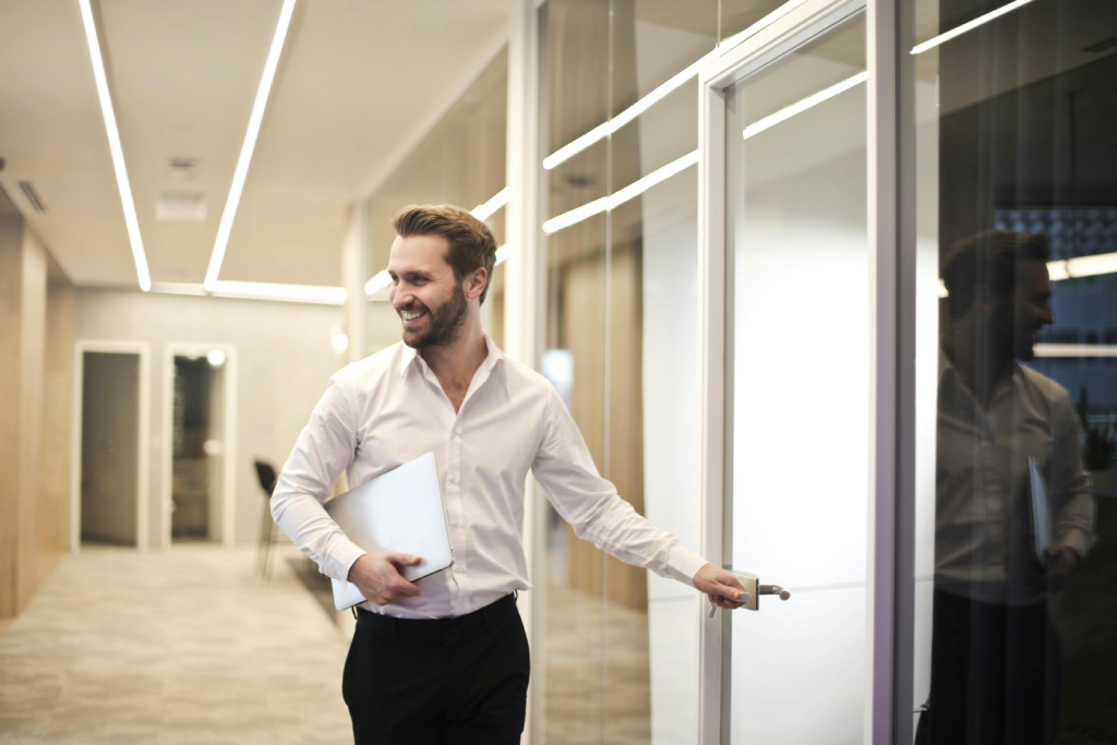 Man in a white shirt and black pants opens a glass office door while smiling and holding a laptop under his arm.