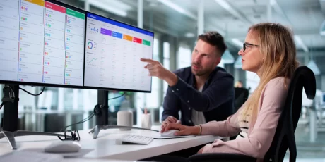 Two colleagues review a data dashboard on dual monitors in a bright open-office setting, with one pointing at the screen.