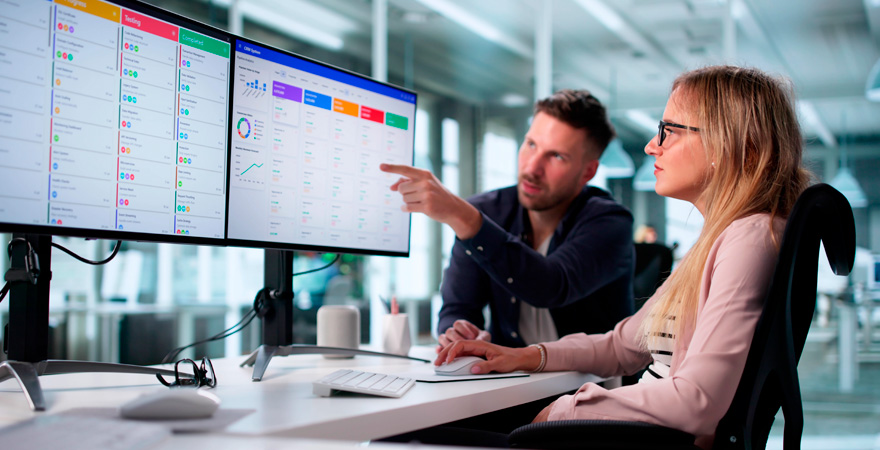 Two colleagues review data dashboards on a dual-monitor setup; one points at the screen.