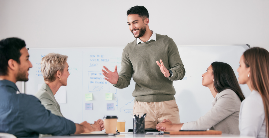 Presenter explains ideas to a diverse team during a meeting, gesturing near a whiteboard with notes on the wall behind them on a conference table backdrop.