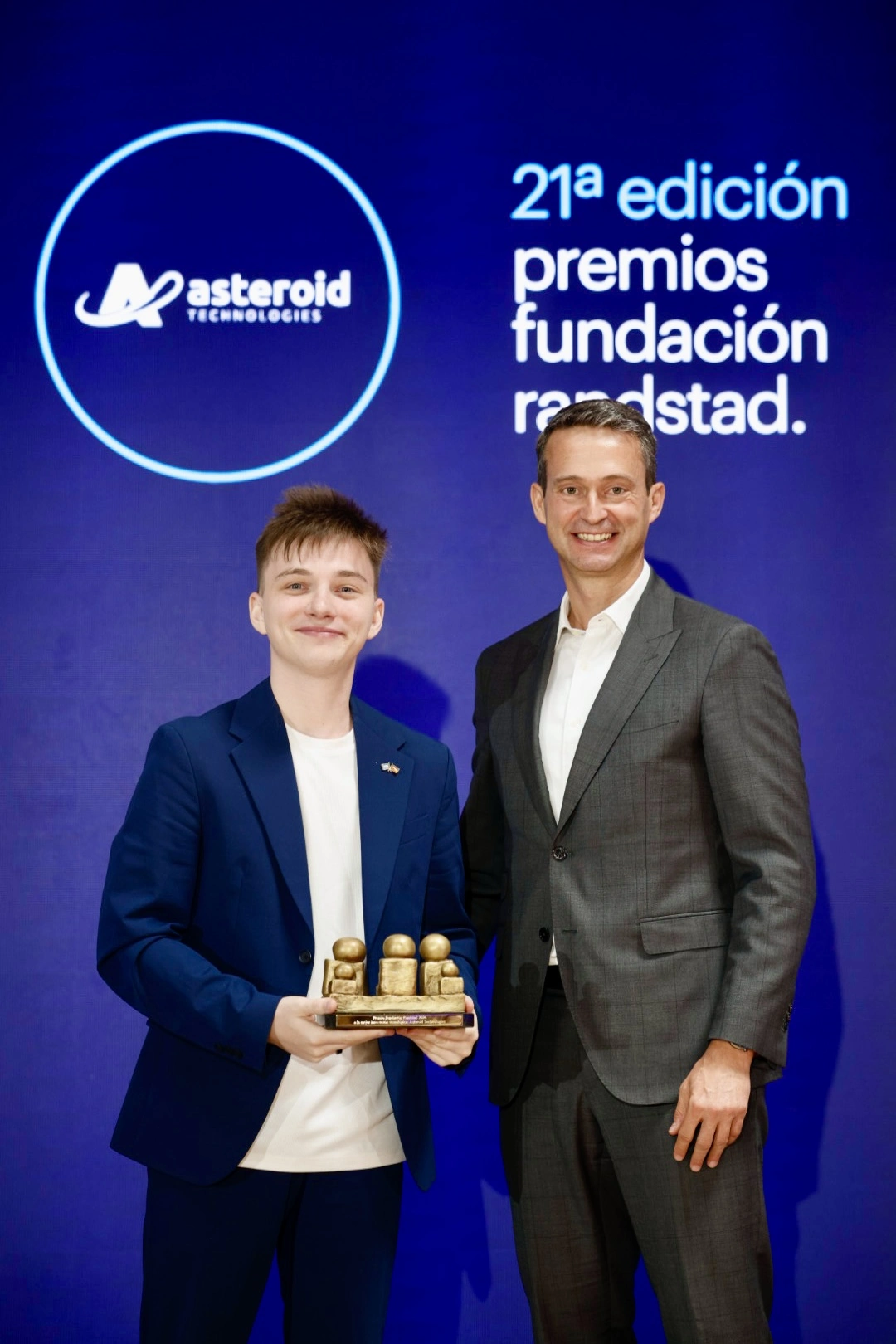 Young man in a blue suit holds a small gold trophy with four figures, standing beside a man in a gray suit at an awards ceremony against a blue backdrop with logos.