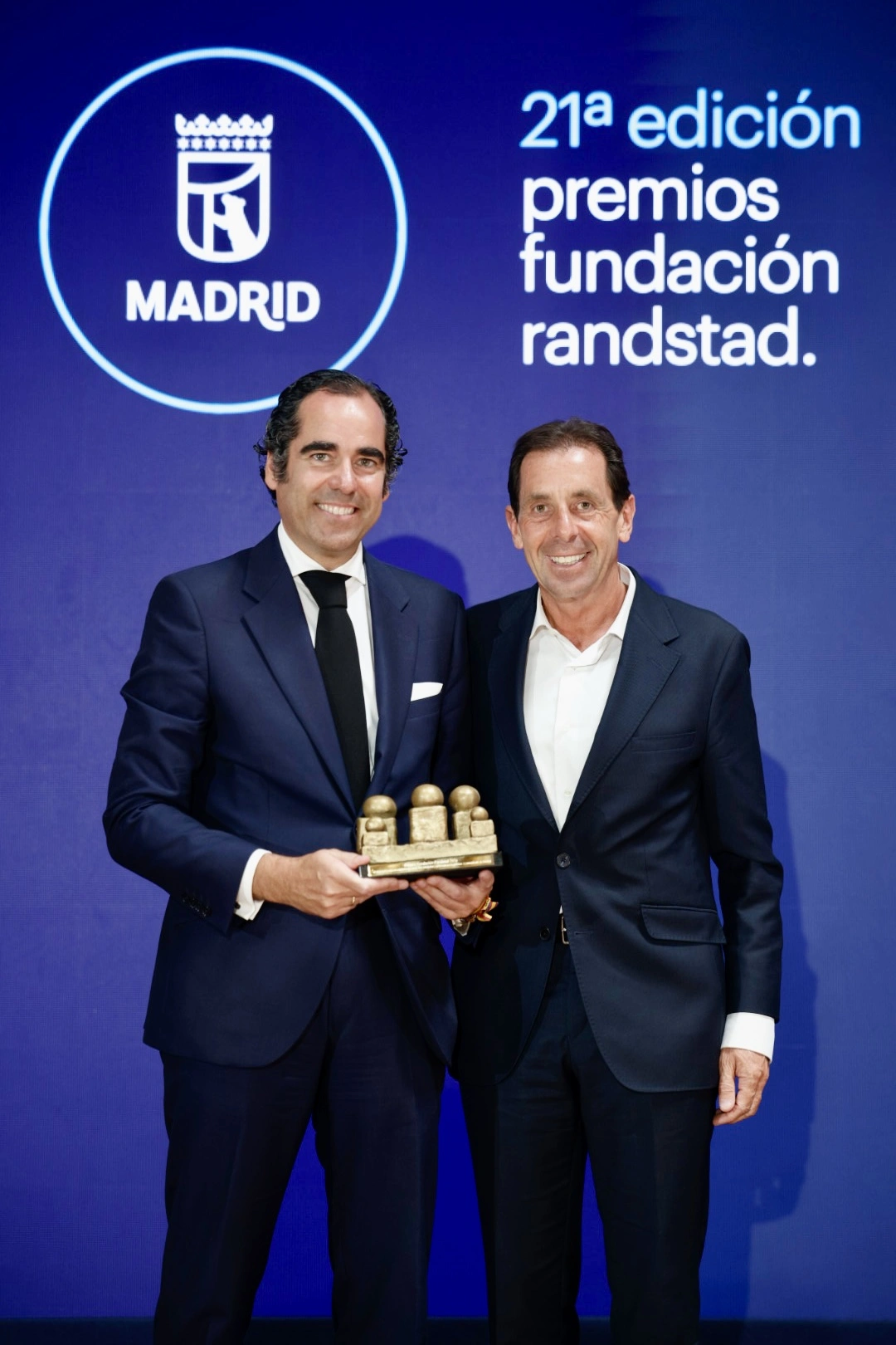 Two men in dark suits pose on stage, holding a gold trophy at an awards ceremony (Madrid logo backdrop).