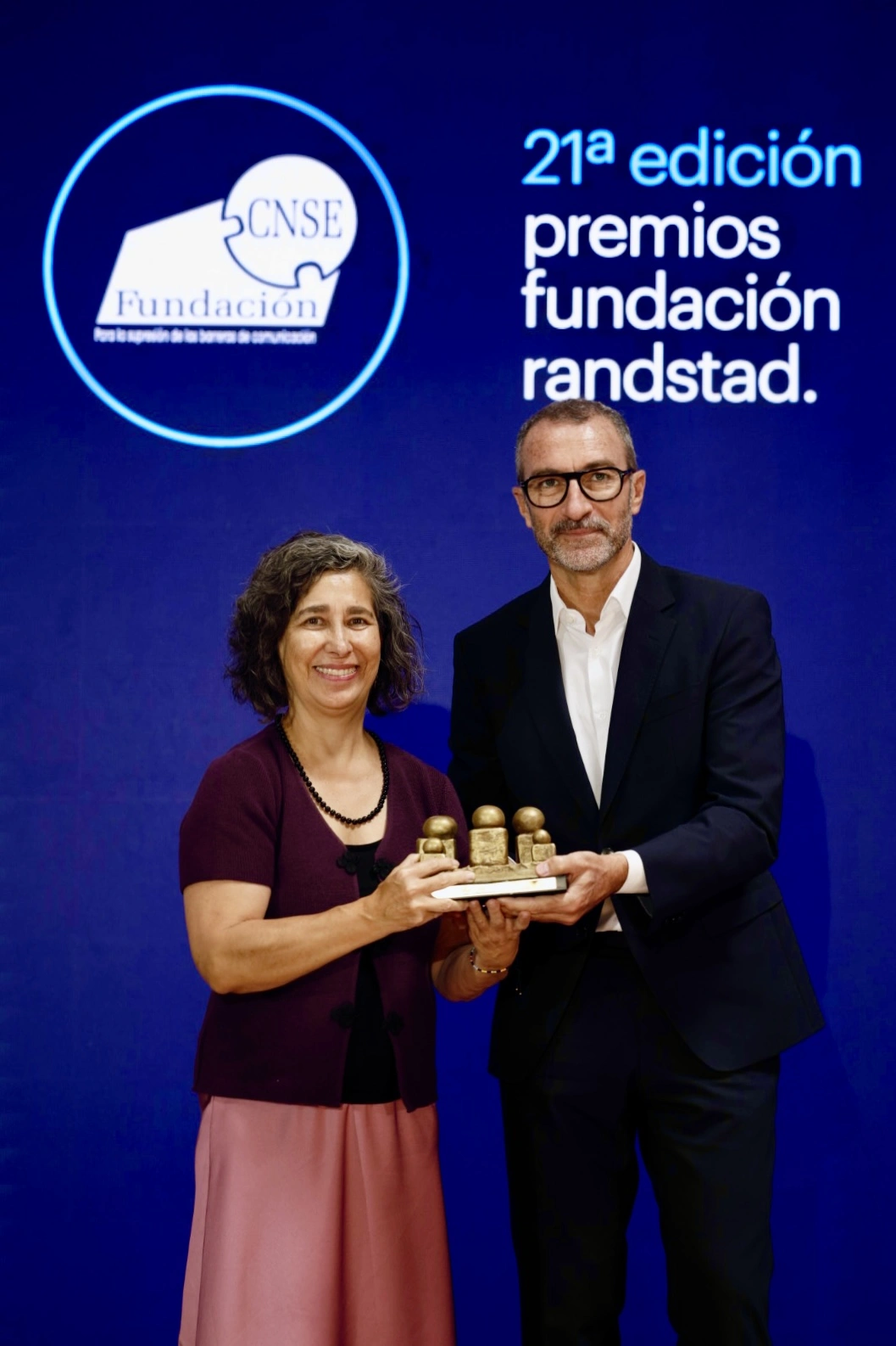 Two adults in front of a blue backdrop holding a small trophy together at an awards event.