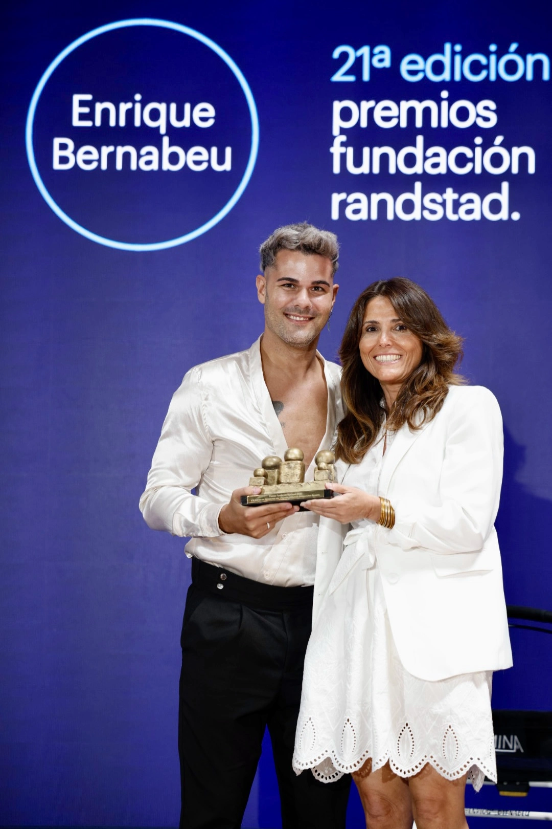 Two people in white outfits smiling while holding a small trophy on a blue stage backdrop with event text.