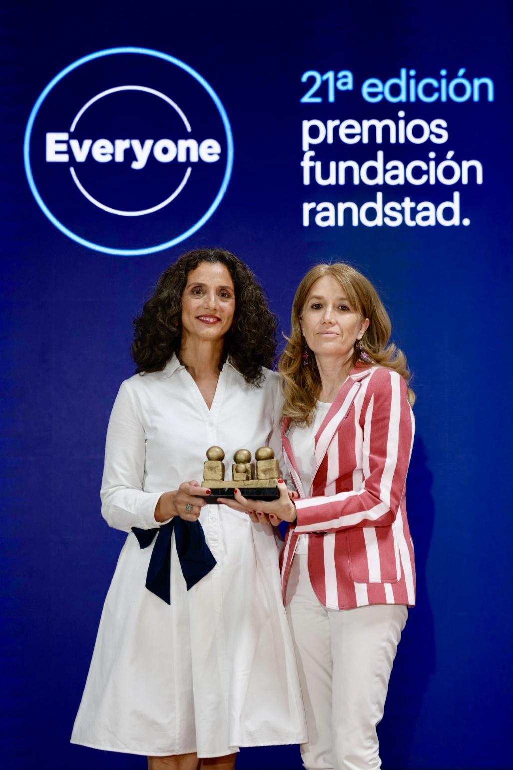 Two women smile while presenting a small trophy at an awards event, blue backdrop with '21a edición premios fundación randstad' and the 'Everyone' logo nearby.