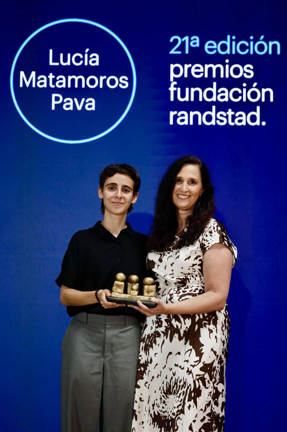 Two women pose with a small trophy at an awards ceremony on a blue backdrop featuring the name Lucía Matamoros Pava and the event text, 21st edition Randstad Foundation awards.
