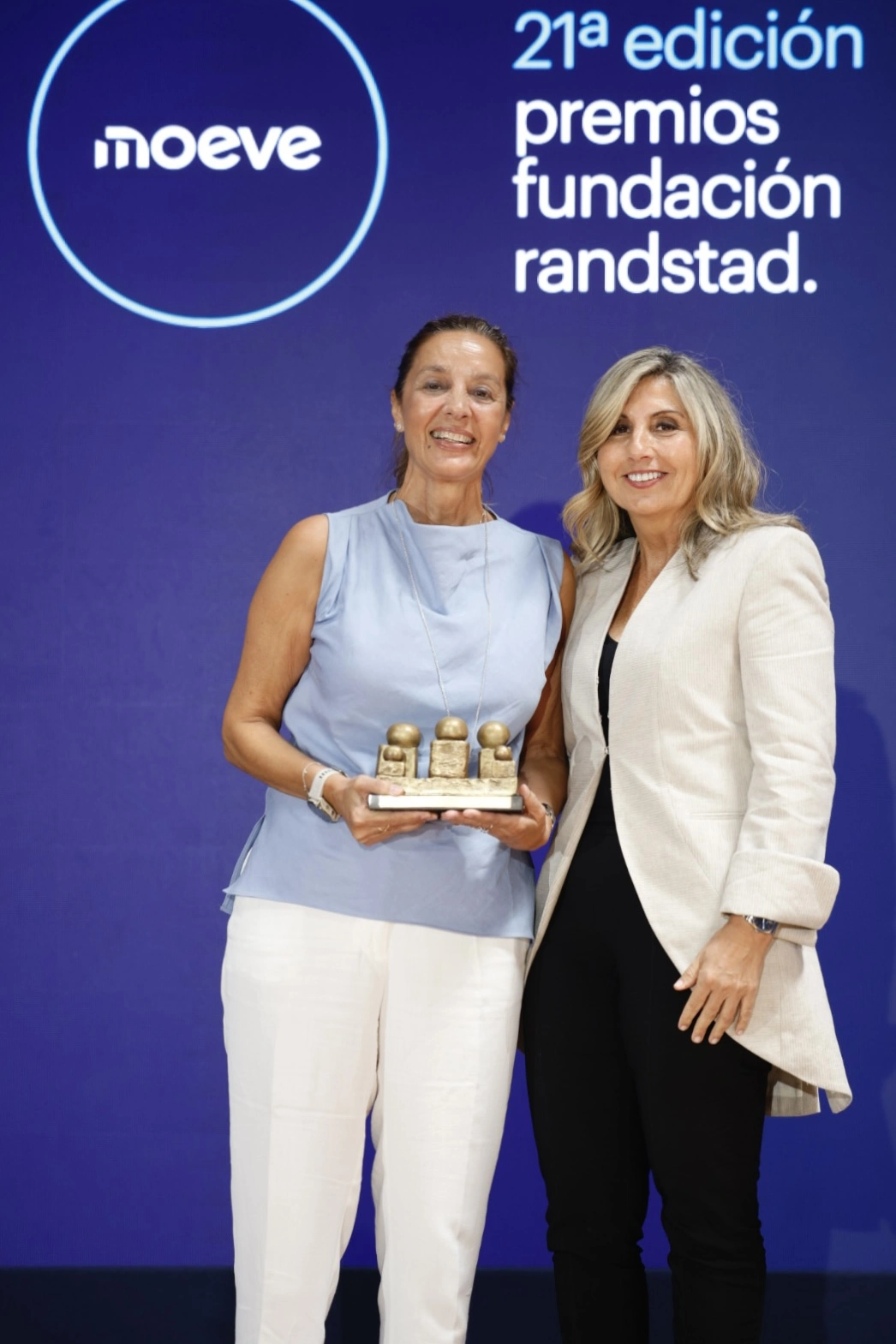 Two smiling women on stage; one holds a small bronze trophy with four figures, as the other stands beside her against a blue backdrop with '21a edición premios fundación randstad' text.