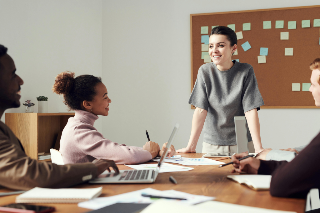 Team meeting: a woman stands at the head of a wooden table, smiling as colleagues work on laptops and papers nearby.