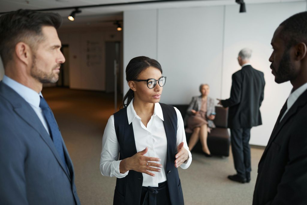 Businesswoman with glasses explains a point to two male colleagues in a modern office lobby or reception area.