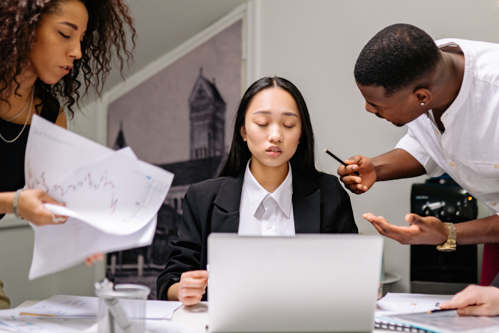 Team of three collaborating around a laptop; the center woman sits with eyes closed while colleagues review papers nearby.