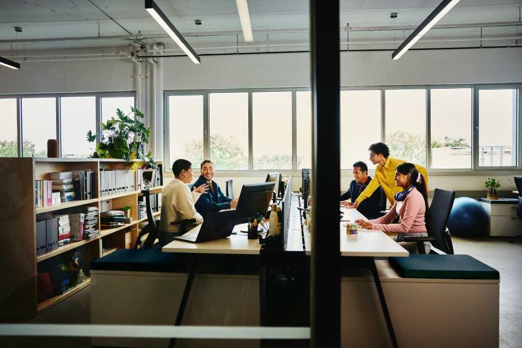 Open-plan office with six coworkers at long desks, laptops open, collaborating near large windows and a dark central divider.
