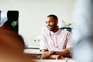Smiling man in a pink patterned shirt sits at a desk in an office setting.