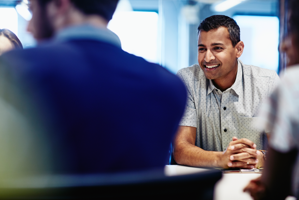 Smiling man in a white dotted shirt talked with colleagues at a meeting table in a bright office setting.
