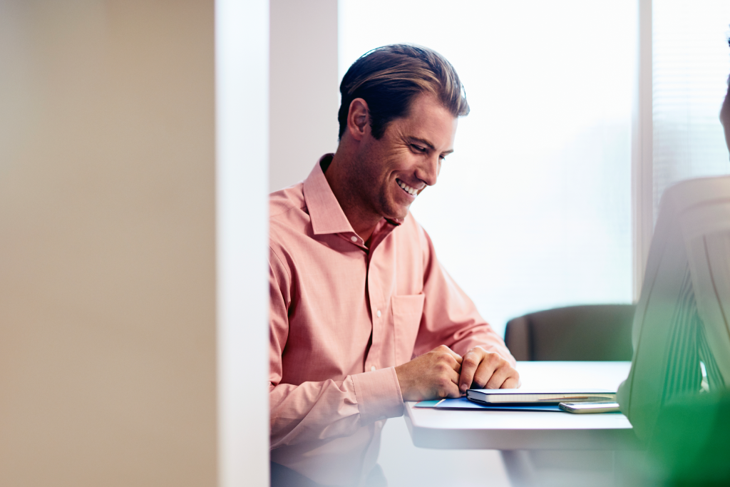 Man in a pink button-down shirt smiles while working at a white desk with a tablet and phone in an office setting.