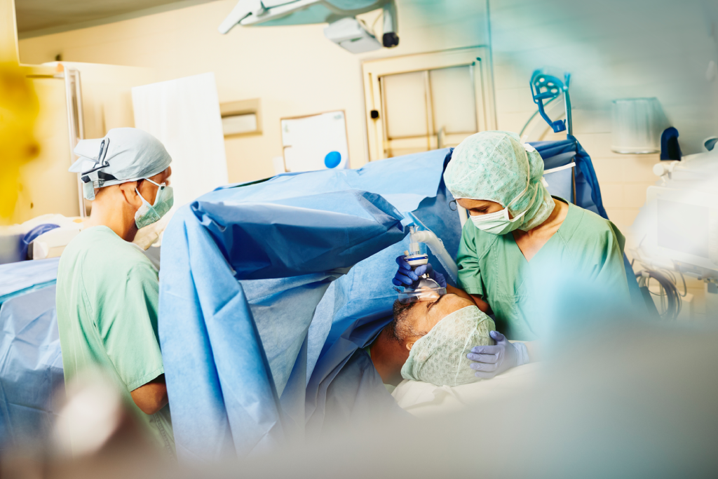 Surgeons perform an operation in a sterile operating room with a patient under anesthesia and blue drapes covering the area.