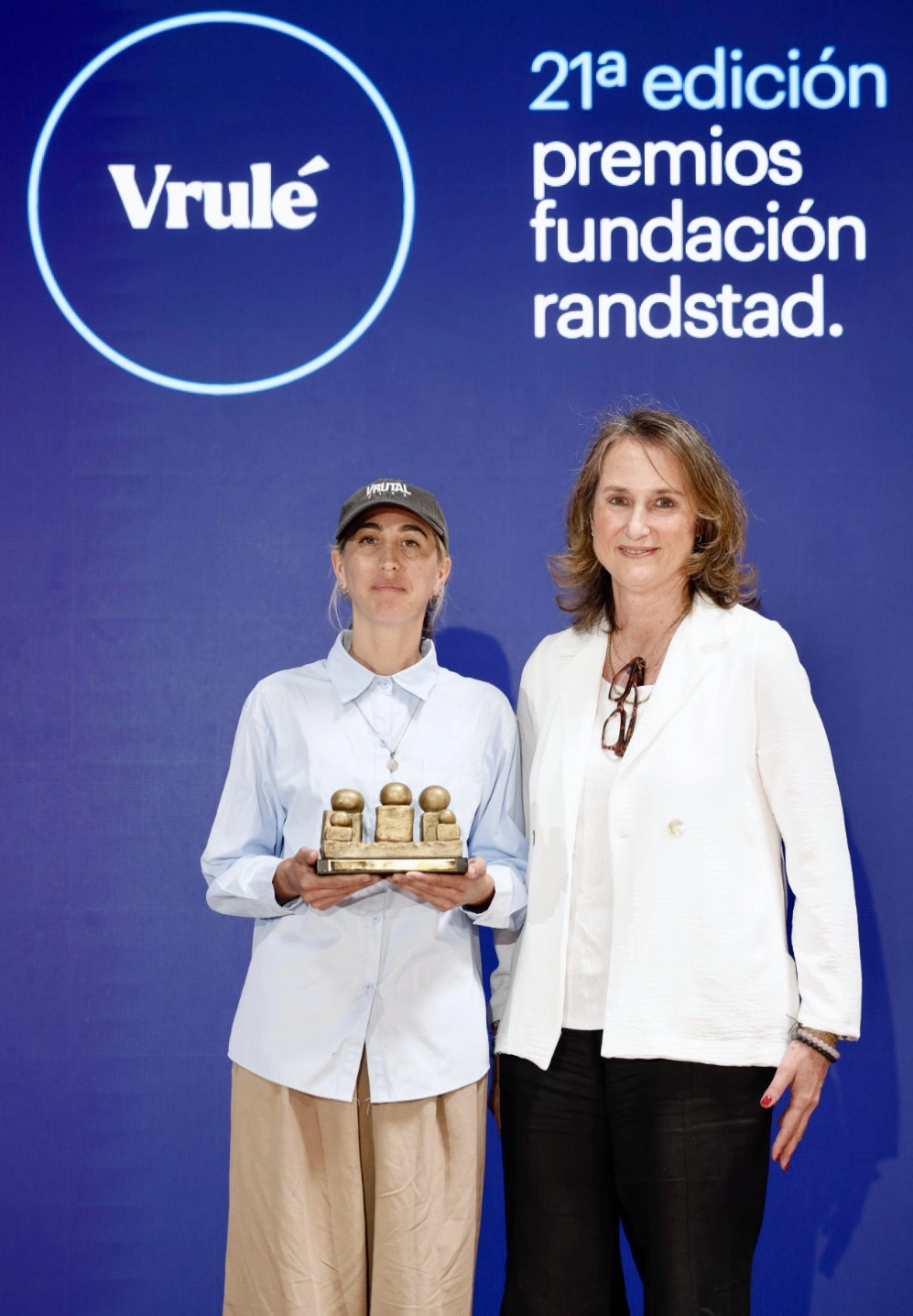 Dos mujeres posan en un escenario frente a un fondo azul con el logo de Vrulé y texto de premios, una de ellas sostiene un trofeo dorado en forma de figuras humanas.