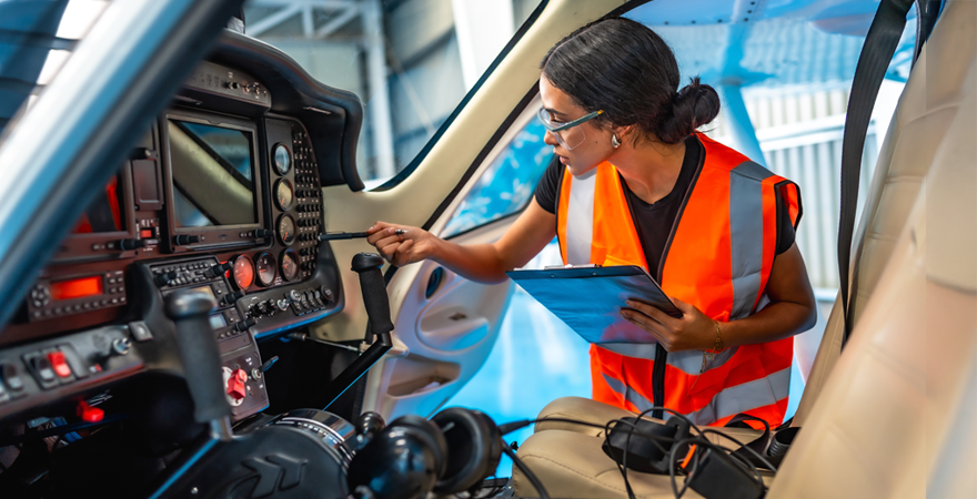 Maintenance technician in an orange safety vest inspecting an aircraft cockpit with a clipboard in hand.