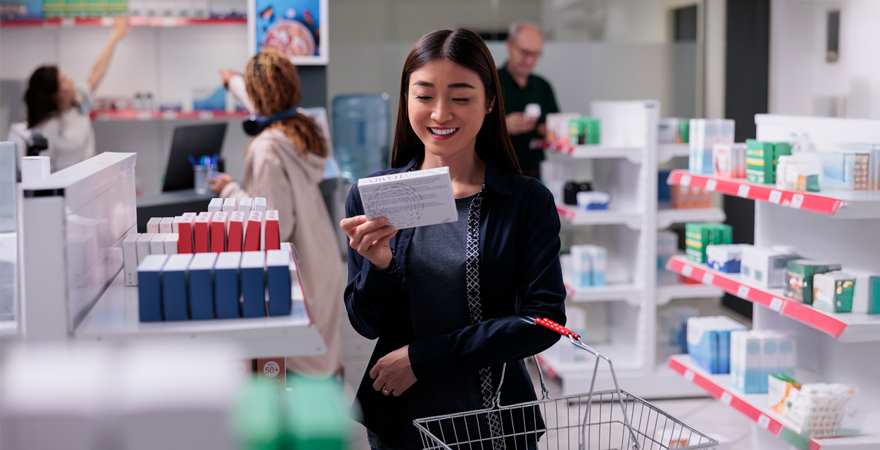 Woman shopping in a pharmacy, reading a card while holding a small shopping cart with medicines on the shelves behind her