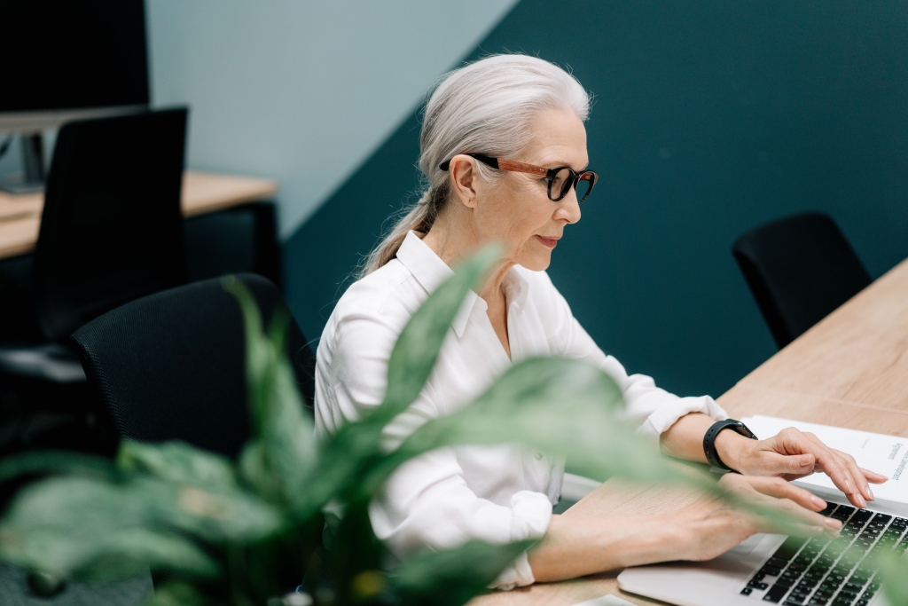 Older woman with gray hair and glasses typing on a laptop at a wooden desk in an office setting.