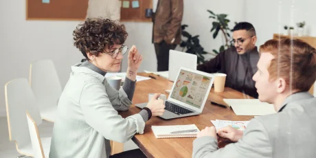 Team members in a modern office sit around a wooden table; woman with curly hair and glasses gestures as charts display on her laptop screen.