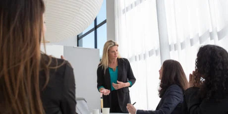 Businesswoman stands at a round table presenting to three colleagues in a bright, modern office.