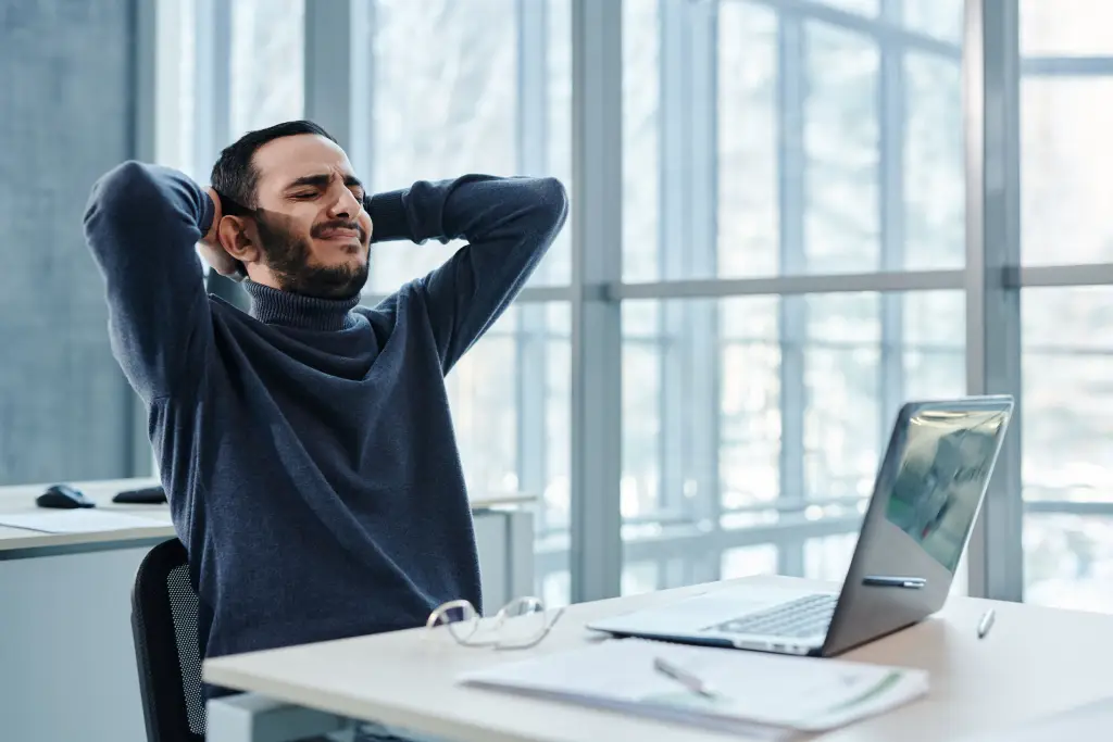 Man in a blue sweater stretches with hands behind his head at a bright office desk, eyes closed.