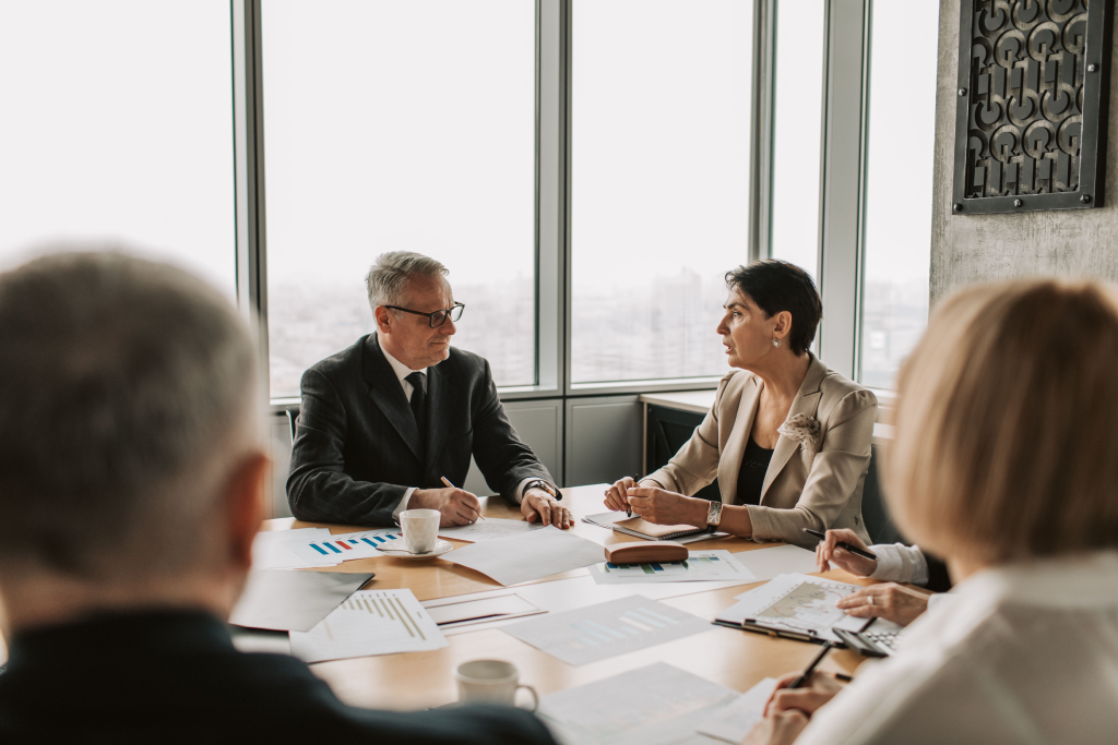 Four professionals in a boardroom discuss documents at a table by large windows with a city view behind them.