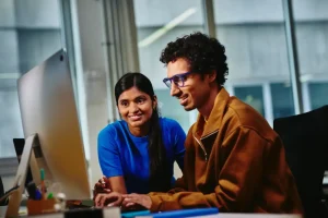 Two colleagues collaborate at a desktop computer in a bright office setting, smiling at the screen the monitor angles left.”,