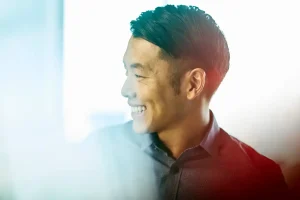 Man with short black hair smiling in profile, facing left, wearing a dark collared shirt.
