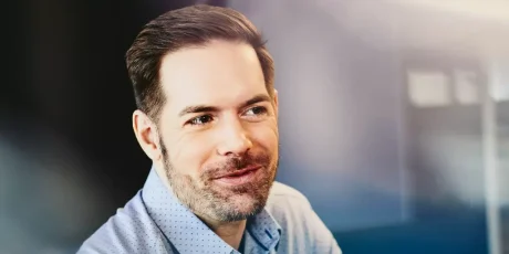 Smiling man in a light blue button-down shirt in an office, looking to the side.