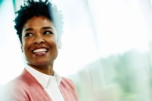 Smiling Black woman in a pink blazer and white shirt indoors, looking off to the side.