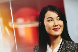 Portrait of a smiling Asian woman with long dark hair, wearing a textured blazer and white blouse, looking to the right.