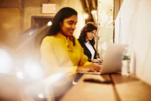 Two women work on laptops at a long shared desk in a warmly lit office space.