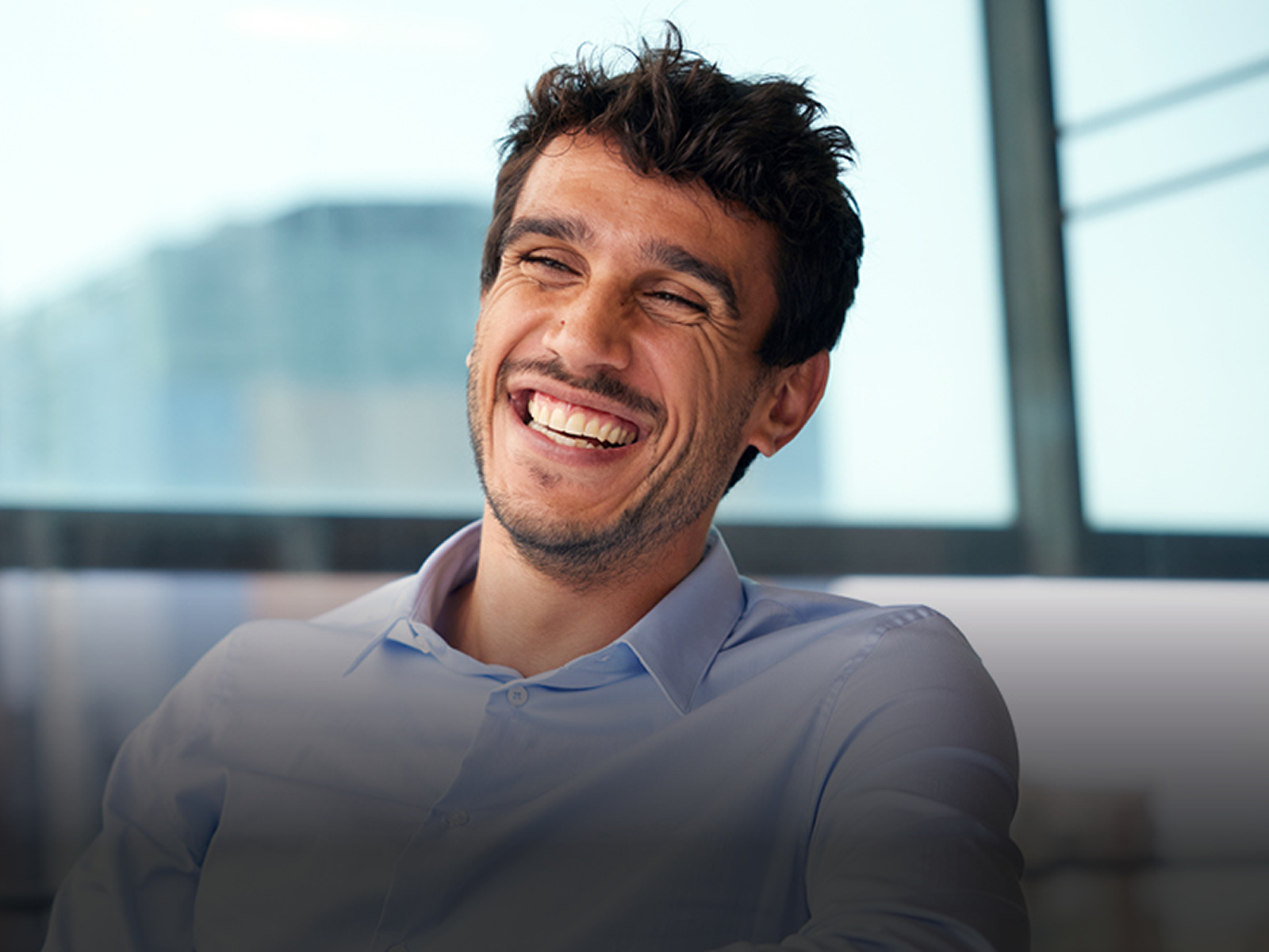 Smiling man with dark hair and stubble, wearing a light blue shirt, in an office setting.